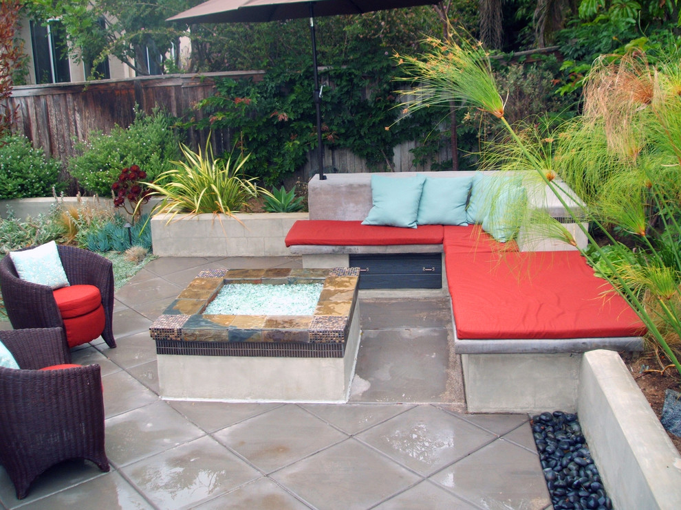 Small concrete patio featuring a built in concrete bench with colorful cushions and accent pillows around a custom natural gas fire pit. Mexican Beach Pebble boarder the ground plane and walls to soften up the space and add textural change. Surrounded by lush green plants, the water feature is the central focal point for this small garden. Modular slate squares transition from the garden area to the seating area.